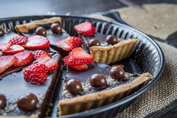 Sliced chocolate tart or pie topped with strawberries, chocolate candy and almonds placed in the tart's pan. Delicious gluten-free homemade crust or tartlet. Side view.