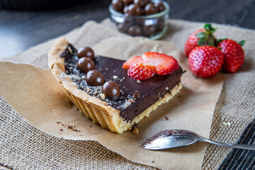 Chocolate tart or pie slice topped with strawberries, chocolate candy and almonds on rustic brown cloth. Dessert spoon in background. Delicious gluten-free homemade crust or tartlet. Back view.