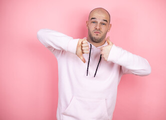 Young bald man wearing casual sweatshirt over pink isolated background Doing thumbs up and down, disagreement and agreement expression. Crazy conflict