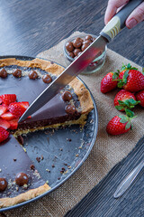 Woman cutting chocolate tart or pie topped with strawberries, chocolate candy, and almonds using a kitchen knife. Delicious gluten-free homemade crust or tartlet. Side view.