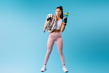 A muscular fitness girl in sport tight leggings holds a rope knot and a protein shake on her shoulder. Posing against a blue background with empty space.