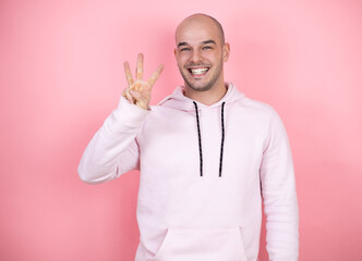 Young bald man wearing casual sweatshirt over pink isolated background showing and pointing up with fingers number three while smiling confident and happy