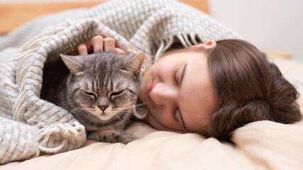 A teenage girl and a British cat lie under a blanket on the bed and sleep. Keeping warm in the cold winter Pets and friendship concept. Quarantine. Holidays. Stay home. Self-isolation. Social distance