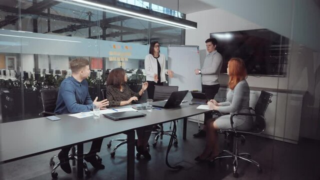 Diverse group of young people having discussion during business meeting in office. Some members of startup team standing by flipchart and reporting, others sitting at table and asking questions
