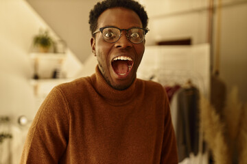 Obraz premium Positive overjoyed young black guy with stubble posing at home with mouth wide opened, exclaiming, cheering at great news, celebrating success or victory, being lucky, having excited look