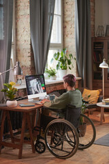 Rear view of disabled freelancer sitting at the table in front of computer monitor and working online at home
