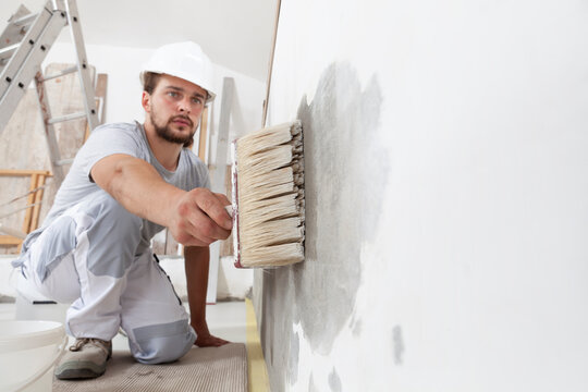 construction worker painter man with protective helmet, brush in hand restores and paint the wall, inside the building site of a house, closeup