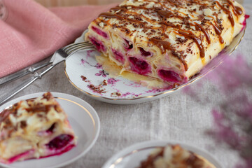 Cake from rolled pancakes with custard, cherries and grated chocolate on white plate, on a light background. Selective focus.