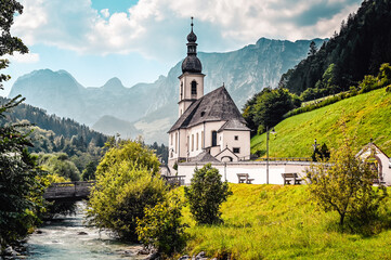 Fototapeta premium Amazing alpine landscape in the Bavarian Alps with the church of St. Sebastian beside a mountain river. The church is standing in a valley surrounded by forested hills and rough mountains.