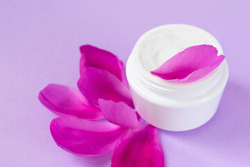 Close up view jar full of white moisturizing cream with peony oil essential or extract and bright petals on pink background