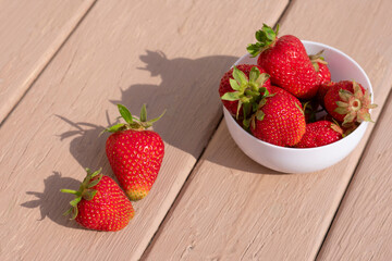 Red ripe strawberries in white bowl. Strawberries on hard summer sunlight with long shadows onwooden background