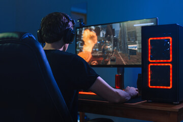 Young professional gamer playing tournaments online video games on computer with headphones in his room, red and blue neon color. Cybersport concept. Back view