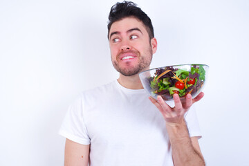 Coquettish Young handsome Caucasian man holding a salad bowl against white background smiling happily, blinking at camera in a playful manner, flirting with you.