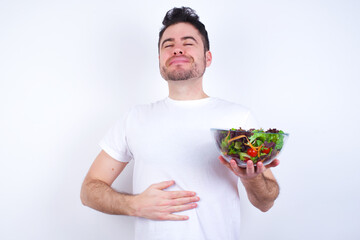 Young handsome Caucasian man holding a salad bowl against white background touches tummy, smiles gently, eating and satisfaction concept.