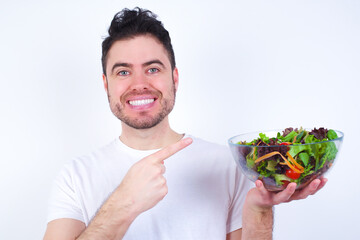 Young handsome Caucasian man holding a salad bowl against white background pointing and holding hand showing adverts