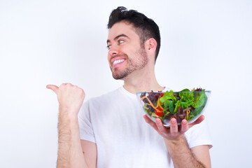 Charming Young handsome Caucasian man holding a salad bowl against white background looking at copy space having advertisements