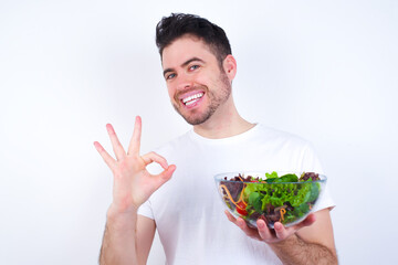 Young handsome Caucasian man holding a salad bowl against white background showing both hands with fingers in OK sign. Approval or recommending concept