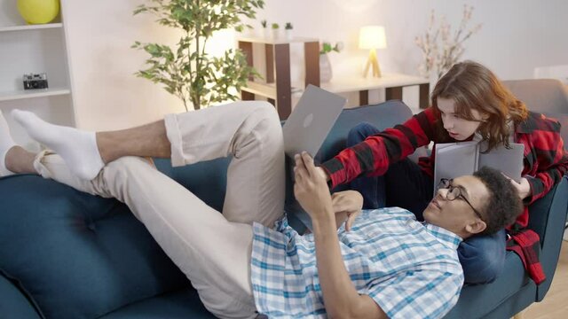 Couple of interracial teen students doing homework on laptop and reading book