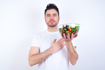 Young handsome Caucasian man holding a salad bowl against white background Swearing with hand on chest and open palm, making a loyalty promise oath