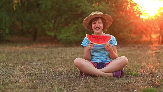 Funny little girl smile and eat watermelon slice sitting on grass with crossed legs in summer park with sunset light.