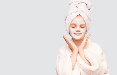 Pleased child girl with wrapped towel on head delighted expression isolated on white background, applies natural beauty product on face, unclogs pores, healthy skin