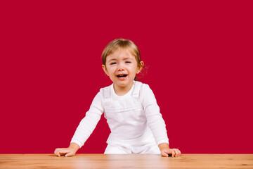 Naklejka premium Blonde toddler girl wearing casual white clothes is scared and crying girl standing over wooden table on red background