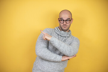 Young bald man wearing glasses over yellow background pointing with hand and finger to the side