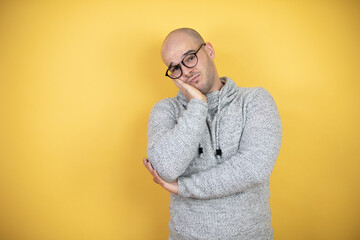 Young bald man wearing glasses over yellow background thinking looking tired and bored with crossed arms