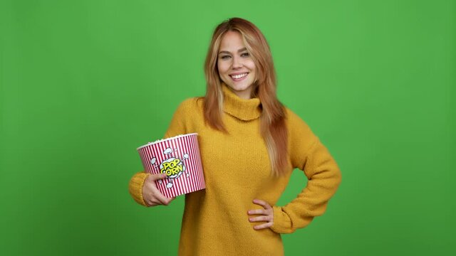 Teenager girl holding a bowl of popcorns posing with arms at hip and laughing over isolated background