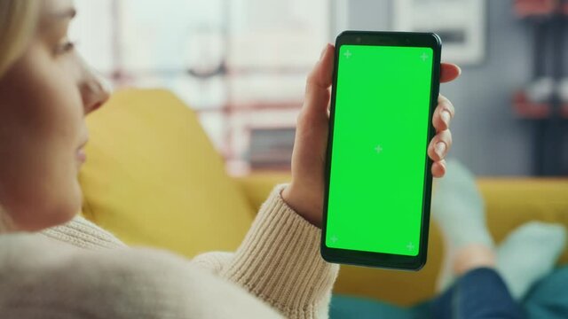 Beautiful Authentic Female Using a Smartphone with Green Screen Mock Up Display at Home Living Room while Lying on a Couch Sofa. She's Browsing the Internet and Checking Videos on Social Networks.