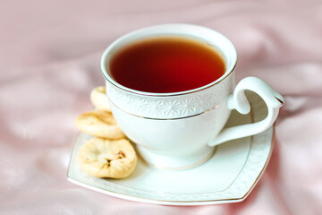 White cup with tea with a saucer, next to dried dates dessert on a silk tablecloth.