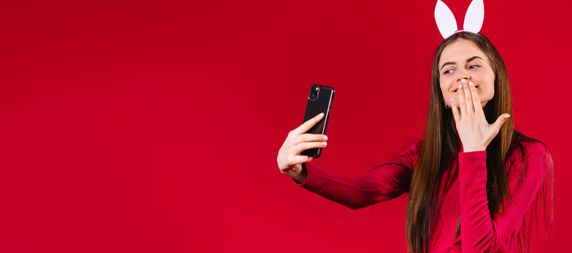 Banner- long format with empty space for Easter advertisement. Beauty blogger wearing bunny white ears, influencer smiling in live stream with followers. Easter atmosphere on red background