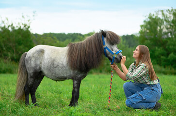 Caucasian young woman and her pony are on the pasture face to face. Female trainer is caressing and...