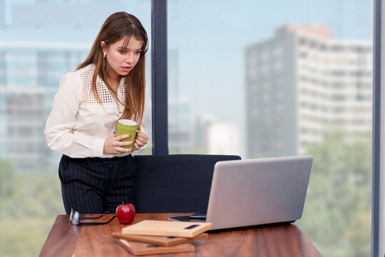 Joven mujer arquitecta con un caf&eacute; en la mano  trabajando desde casa, mirando su computadora con cara de sorpresa. concepto de trabajando en casa