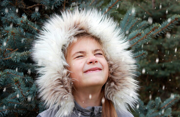 a little girl stands under the flying snow against the background of fir branches