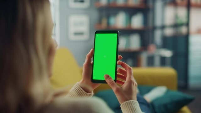 Beautiful Authentic Female Using a Smartphone with Green Screen Mock Up Display at Home Living Room while Lying on a Couch Sofa. She's Browsing the Internet and Checking Videos on Social Networks.
