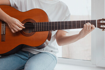 Fototapeta na wymiar Cropped shot of young woman in the white t-shirt and blue jeans is sitting on the windowsill and playing acoustic guitar. Close up of the hands. Girl picks a chord clamping frets on the fretboard