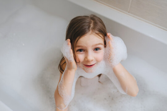 A small, smiling, beautiful red-haired girl with long hair, a child bathes, washes her face with foam in a white bath. Cheerful photography.