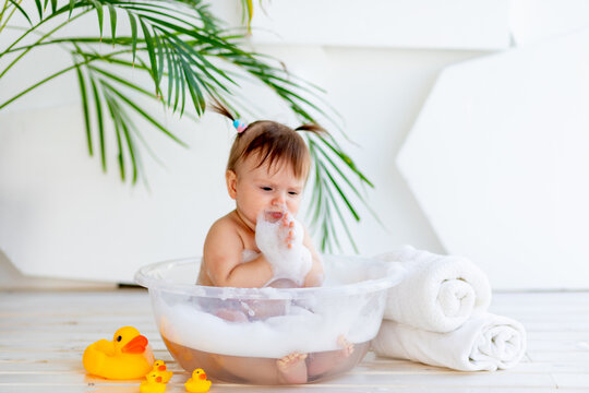 funny little child girl sitting in a basin with foam and water in a bright room at home and trying the foam taste