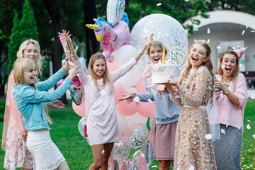 Six girls have fun at a birthday party in the park. They're wearing pink dresses and a bunch of pink balloons. They have cake and firecrackers