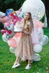 Portrait of a girl with a cake in the park. She's wearing a pink dress and carrying a bunch of giant balloons.