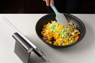 Woman putting vegetables into frying pot and watch virtual culinary class