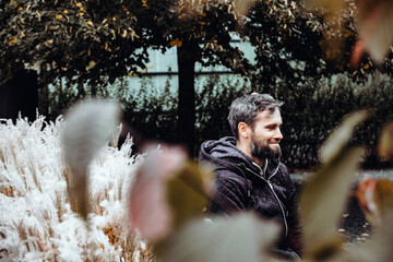 Handsome middle-aged Caucasian man portraited in the city, sitting on a bench with grey and black hair having a friendly look on his face, wearing a blue jacket and jeans. Fall colourful background.