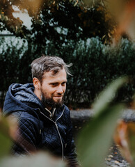 Handsome middle-aged Caucasian man portraited in the city, sitting on a bench with grey and black hair having a friendly look on his face, wearing a blue jacket and jeans. Fall colourful background.