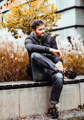 Handsome middle-aged Caucasian man portraited in the city, sitting on a bench with grey and black hair having a friendly look on his face, wearing a blue jacket and jeans. Fall colourful background.