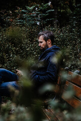 Handsome middle-aged Caucasian bearded man portrayed in the city, sitting on a park bench, holding a cell phone, with grey and black hair having a friendly look on his face, wearing city clothes.