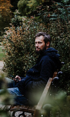 Handsome middle-aged Caucasian bearded man portrayed in the city, sitting on a park bench, holding a cell phone, with grey and black hair having a friendly look on his face, wearing city clothes.