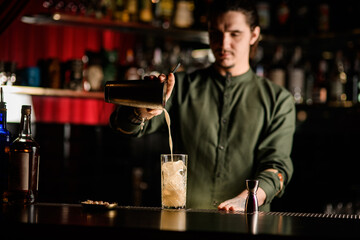 professional bartender pours cocktail from steel shaker into crystal glass