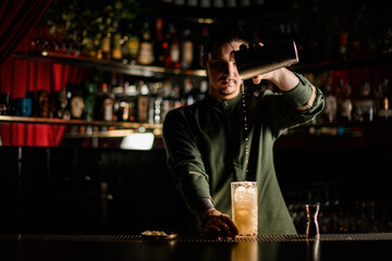view of young bartender pouring cocktail from steel shaker into glass in dark bar