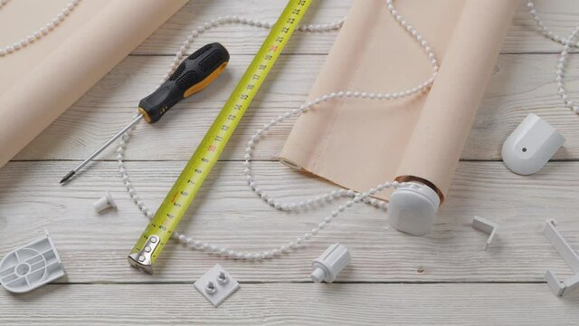 The two beige roller blinds, spare parts, mesure tape and screwdriver on a white wooden table. Preparation for the installation or repair of roller blinds.